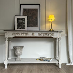 Könnte beinhalten: Ein weiß-grauer Konsolentisch mit verzierten Schnitzereien und einer grauen Platte. Der Tisch hat ein unteres Regal mit einer dekorativen Urne und einem Buch. Ein gerahmter Druck und eine Lampe mit gelbem Schirm stehen oben.