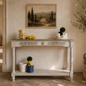 May include: A rectangular wooden console table with a distressed white finish, featuring carved floral details on the front drawer panel, fluted legs, and a lower shelf. The table is topped with a glass jar of lemons and decorative candles.