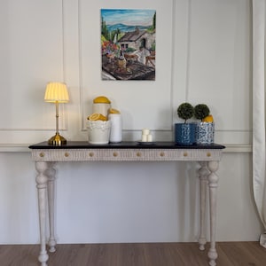 Puede incluir: Una consola decorativa con tapa negra y patas blancas. La mesa está adornada con una lámpara dorada, una pintura y objetos decorativos, incluyendo limones, pequeñas plantas en macetas y velas. La mesa mide 122 cm de largo.
