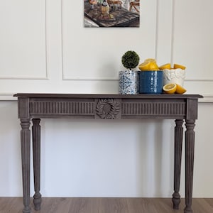 Puede incluir: Una consola de madera gris con patas estriadas y un diseño de hojas talladas. La mesa sostiene tres macetas decorativas, una con un pequeño topiario, una azul y otra blanca, llenas de limones. Una obra de arte de una escena europea cuelga encima.