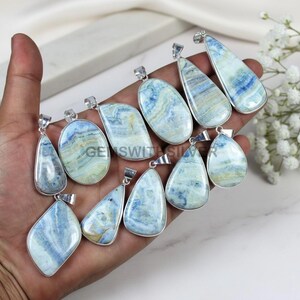 May include: A collection of gemstone pendants in various shapes, including teardrop, oval, and triangular. The pendants feature a blue and white marbled pattern and are set in silver-colored metal. The pendants are displayed on a hand.