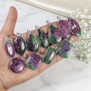 May include: A selection of gemstone pendants in diverse shapes, such as oval, teardrop, and rectangular. The pendants display a blend of deep red and green hues, set in silver-toned metal. The gemstones are held in a hand.
