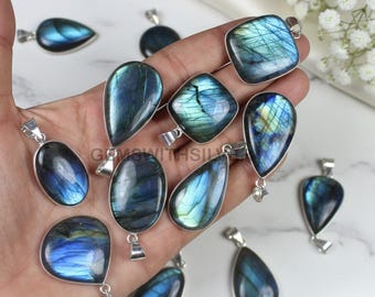 Può includere: Una collezione di pendenti in labradorite di varie forme, tra cui a goccia, ovale e quadrata. Ogni pendente presenta un'ansa in argento e mostra sfumature iridescenti blu e verdi. I pendenti sono esposti su una superficie chiara.