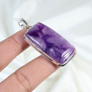 Puede incluir: Un colgante rectangular de amatista con un bisel plateado. La piedra púrpura pulida tiene vetas más claras y oscuras. El colgante tiene un pequeño lazo en la parte superior para una cadena.