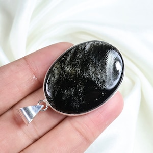 May include: Oval-shaped pendant with a black and gold gemstone set in a silver-colored metal frame. The gemstone has a reflective, polished surface with a unique pattern. The pendant has a silver bail for a necklace.