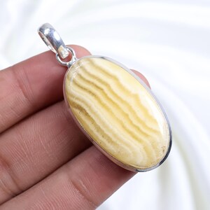 Puede incluir: Colgante ovalado con una piedra a rayas crema y beige engastada en un marco de metal plateado. El colgante tiene un lazo en la parte superior para una cadena. La piedra tiene un acabado pulido.