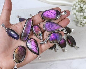 Può includere: Una collezione di pendenti in labradorite lucida di varie forme, tra cui ovali, a goccia e oblunghe. Ogni pendente presenta una vibrante esposizione di colori viola e iridescenti, incastonati in una montatura in metallo argentato. I pendenti sono esposti in una mano.