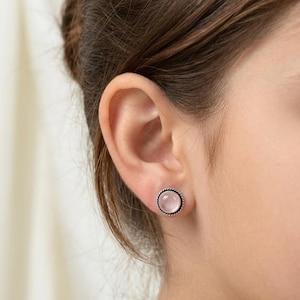 May include: A round stud earring featuring a smooth, pale pink cabochon stone set in a silver-toned metal frame with a twisted rope-style border. The earring is secured to the earlobe, showcasing a simple, circular design with a polished finish.