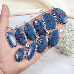 May include: A collection of oval-shaped pendants with a silver-colored border. The pendants feature a blue-green stone with red and purple speckles. The pendants are displayed in a hand.