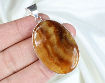 May include: An oval-shaped pendant featuring a polished, translucent brown and amber-coloured stone with natural banding patterns, encased in a smooth silver-toned metal setting with a triangular bail at the top for attachment to a chain.