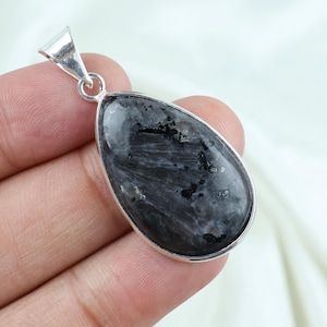 May include: A teardrop-shaped pendant featuring a polished, dark grey stone with black inclusions and a shimmering, fibrous texture. The stone is encased in a smooth, silver-toned metal setting with a triangular bail at the top for hanging.