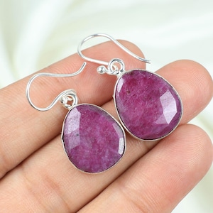 May include: A pair of silver earrings featuring large, faceted, oval-shaped, deep pink gemstones. The gemstones are set in a silver bezel and suspended from simple silver ear wires. The earrings are displayed against a soft, white background.