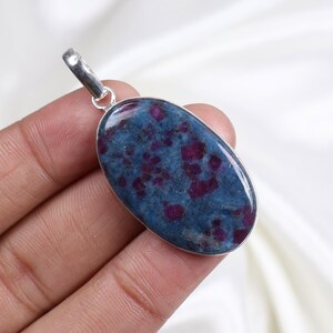 Può includere: Ciondolo ovale con anello d'argento. Il ciondolo presenta una pietra blu con inclusioni rosse. La pietra è incastonata in una lunetta d'argento. Il ciondolo è tenuto in una mano.