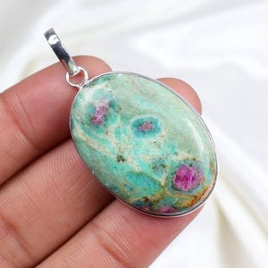 Puede incluir: Un colgante de forma ovalada con una piedra preciosa pulida de varios colores, con una base verde menta dominante, vetas blancas veteadas e inclusiones de color magenta rosado, engastado en un marco de metal plateado liso y pulido.