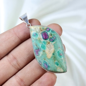 Puede incluir: Un colgante en forma de marquesa con una piedra de Anyolita pulida que presenta una matriz de zoisita verde vibrante e inclusiones de rubí magenta, engastado en un marco de metal plateado con una anilla triangular en la parte superior.