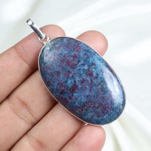 Puede incluir: Colgante ovalado con una argolla plateada. El colgante presenta una piedra pulida con una mezcla de tonos azules y granates. La piedra está engastada en un bisel plateado.