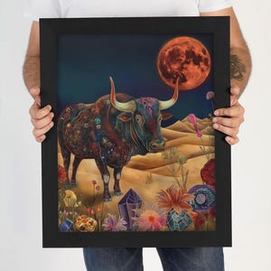 Op de afbeelding: Ingelijste kunstafdruk met een kleurrijke illustratie van een stier tegen een woestijnlandschap met een grote rode maan op de achtergrond. De stier is versierd met ingewikkelde patronen en omgeven door bloemen en kristallen.