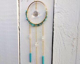 May include: A gold-toned dreamcatcher with turquoise stones and a crystal prism hanging from it. The dreamcatcher is hanging on a white wooden wall.