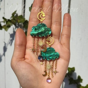 Puede incluir: Pendientes con temática celestial. Presentan colgantes verdes en forma de nube con soles, lunas y estrellas doradas. Los pendientes cuelgan de ganchos y cadenas doradas, con un cristal púrpura en la base. Un accesorio único.