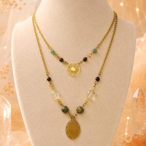 May include: A gold-colored, layered necklace with a sunburst pendant and gemstone accents. The necklace features a mix of clear, green, and maroon-colored beads. The lower layer has an oval-shaped, yellow-toned pendant.