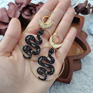 May include: A pair of gold hoop earrings with a crescent moon and snake charm. The snake is black with a white glitter design of stars and moons.