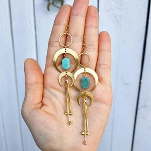 May include: A pair of gold-toned dangle earrings featuring a circular hoop at the top, a crescent moon shape, and a rectangular turquoise-colored stone. Below the stone, a second crescent moon hangs with three thin, dangling gold-toned chains.