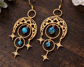 May include: A pair of gold-toned dangle earrings featuring a crescent moon cutout design, dangling star charms, and two round blue beads suspended within circular frames. The earrings hang from simple gold-toned hooks for pierced ears.