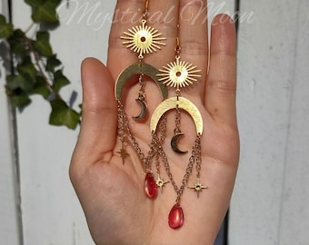 May include: Gold earrings with a sun and moon design. The earrings feature a crescent moon with a small star hanging from it, and a sun with a starburst design. The earrings are accented with red glass beads.