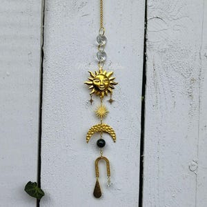 Puede incluir: Encanto de sol, luna y estrella de oro colgando de una cadena con cuentas de vidrio transparentes. El sol tiene una cara y la luna tiene forma de media luna. Una cuenta verde oscuro cuelga entre la luna y un arco dorado.