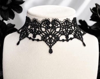 May include: A black lace choker necklace featuring an intricate, symmetrical pattern of swirling filigree and heart-shaped motifs. The design includes delicate scalloped edges and small, dangling teardrop-shaped accents along the bottom border.