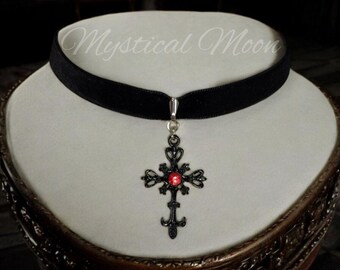 May include: Black velvet choker with a silver pendant featuring a black cross with a red gemstone in the center. The choker is on a white display stand. Mystical Moon is written in white text on the stand.