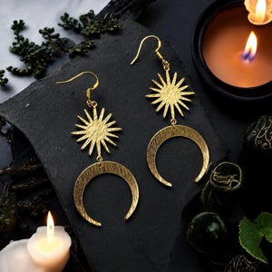 May include: A pair of gold-toned dangle earrings featuring a sunburst charm suspended above a crescent moon charm. Each earring is attached to a standard hook wire. The metal surfaces have a textured, brushed finish throughout the design.