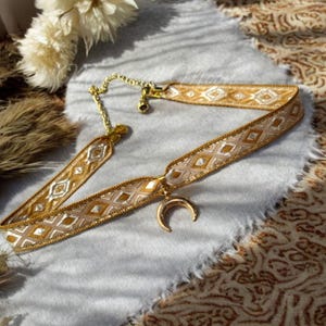 May include: A gold choker necklace with a crescent moon pendant. The choker is made of a gold ribbon with a geometric pattern.