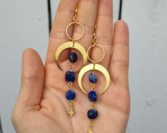 May include: Gold-tone earrings featuring crescent moon shapes, circular accents, and dangling blue lapis lazuli stones. Each earring has a small star charm at the bottom. The earrings are displayed against a neutral background.