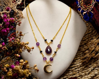 May include: A double-layered gold chain necklace featuring a faceted purple teardrop pendant on the shorter chain and a gold crescent moon charm on the longer chain, accented with small purple beads and tiny black beads along the gold links.