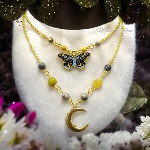 May include: A gold chain necklace with a crescent moon pendant and a butterfly charm. The necklace is made with green and yellow beads. The butterfly charm has a black and blue design with gold accents. The text "Mystical Moon" is visible in the background.