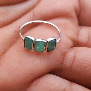 Könnte beinhalten: Ein Silberring mit drei rechteckigen, smaragdgrünen Edelsteinen. Der Ring ist auf einem dünnen Band gefasst und wird auf einer Hand präsentiert, wodurch das Design und die Farbe des Rings hervorgehoben werden.