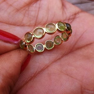 K&ouml;nnte beinhalten: Ein goldener Ring mit einer Reihe von runden, hellgr&uuml;nen Edelsteinen. Der Ring wird in einer Hand gehalten. Die Edelsteine sind gleichm&auml;&szlig;ig um das Band angeordnet und bilden ein durchgehendes Muster.