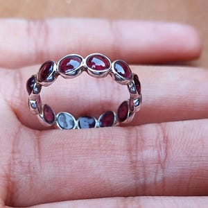 May include: A silver ring with a series of oval-shaped, deep red gemstones set around the band. The gemstones are evenly spaced, creating a continuous pattern. The ring is displayed on a hand.