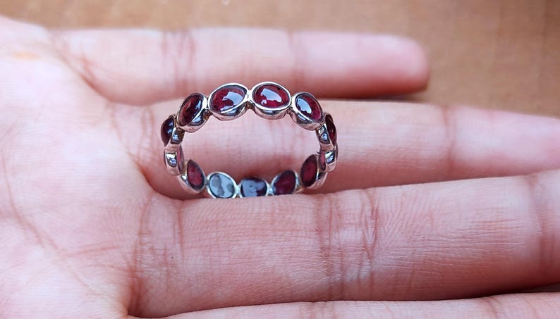May include: A silver ring with a series of oval-shaped, deep red gemstones set around the band. The gemstones are evenly spaced, creating a continuous pattern. The ring is displayed on a hand.