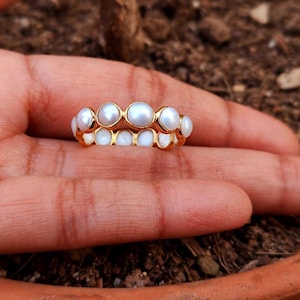 Könnte beinhalten: Ein goldener Ring mit einem durchgehenden Band aus kleinen, runden, weißen Perlen in einer Reihe. Die Perlen sind glatt und glänzend und werden von einem dünnen Goldmetallrahmen gehalten, der jede einzelne Perle umschließt.