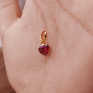 May include: A small heart-shaped pendant featuring a deep red faceted gemstone set in a polished gold-toned metal frame with a matching gold-toned bail at the top for attachment to a chain or necklace, showcasing a classic and elegant design.