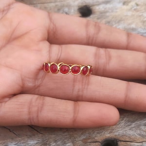 May include: A gold-colored ring with a series of oval-shaped, red gemstones set in a row. The ring is displayed on a hand, showcasing the jewelry's design and color. The gemstones are evenly spaced, creating a continuous pattern.