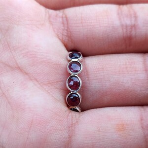 May include: A silver ring with five oval, deep red gemstones. The ring is shown on a person's finger, with the gemstones set in a row. The ring is a simple, elegant design.