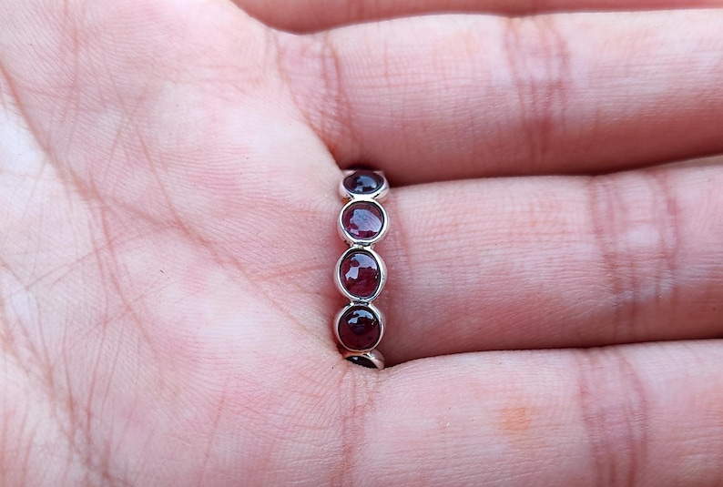 May include: A silver ring with five oval, deep red gemstones. The ring is shown on a person's finger, with the gemstones set in a row. The ring is a simple, elegant design.