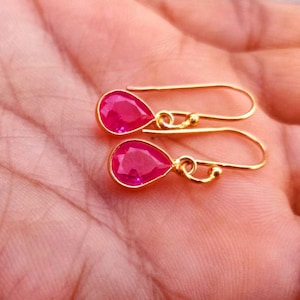 May include: A pair of gold-toned drop earrings featuring faceted, teardrop-shaped hot pink stones. Each earring is suspended from a simple curved gold-toned hook wire, showcasing a polished metal setting that frames the vibrant pink gemstone.