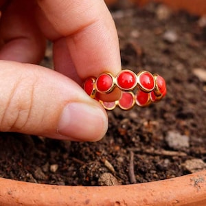 May include: A gold-toned ring featuring a continuous band of small, round, vibrant red cabochon stones set in a bezel style. The ring is held up, showcasing the circular arrangement of the red stones against the metallic gold setting.