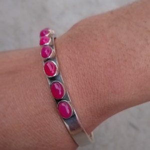 May include: A silver bangle bracelet with pink oval-shaped gemstones set in the metal. The bracelet is open and adjustable.