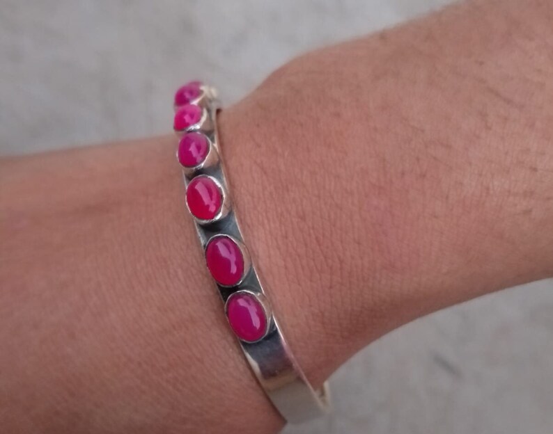 May include: A silver bangle bracelet with pink oval-shaped gemstones set in the metal. The bracelet is open and adjustable.