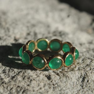 Könnte beinhalten: Ein goldener Ring mit einer durchgehenden Reihe runder, im Cabochon-Schliff gehaltener grüner Edelsteine, die in einzelnen Metallfassungen sitzen. Der Ring zeigt eine Reihe leuchtender, durchscheinender grüner Steine in kreisförmiger Anordnung.